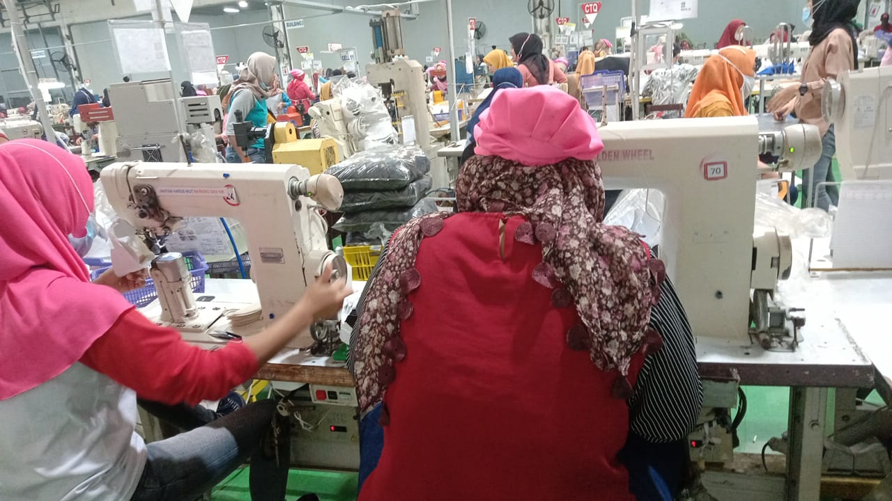 Two women garment workers wearing pink and white uniforms with headwear, their backs facing the viewer. They are stitching cloth in a garment factory in Indonesia. They are surrounded by other garment workers, all women. 