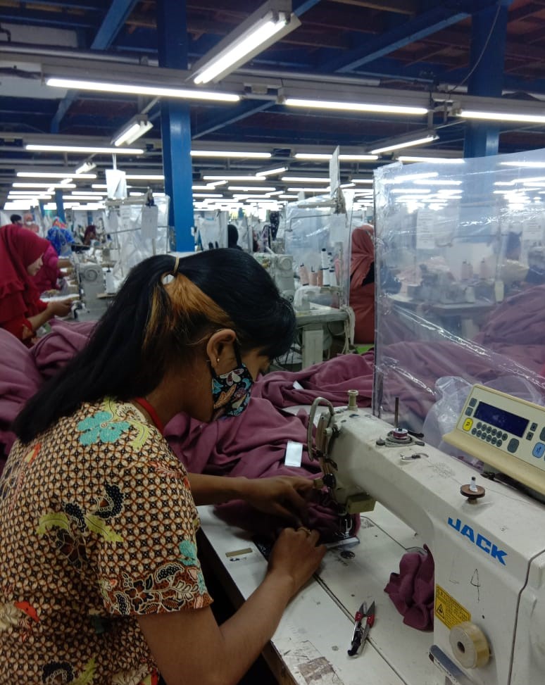 An Indonesian woman with a ponytail is busy at work using her sewing machine to stitch a roll of lavender cloth. She is sitting near two other garment workers in a garment factory. 